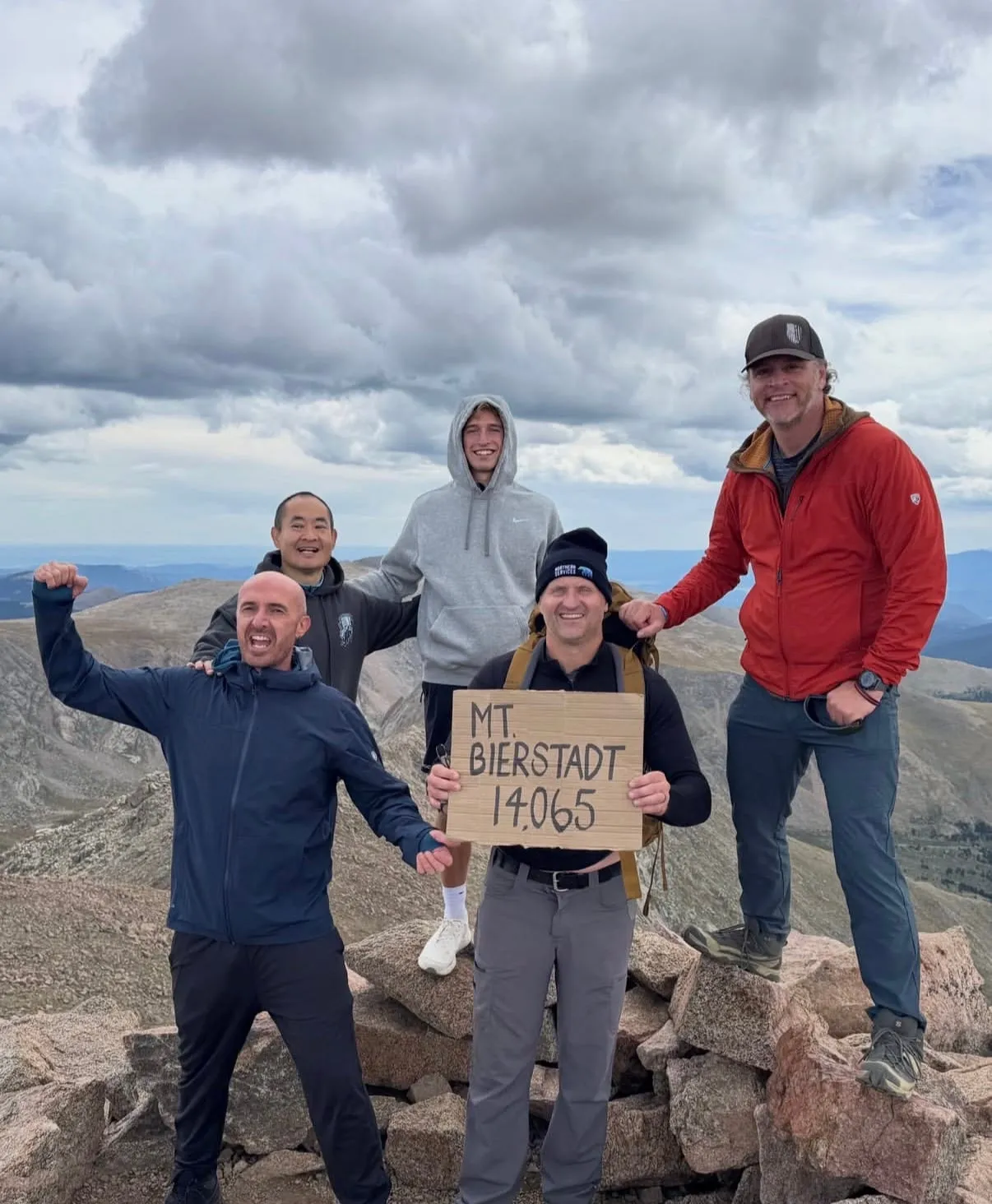 Five hikers on the summit of Mt Bierstadt holding a cardboard sign reading Mt. Bierstadt 14,065