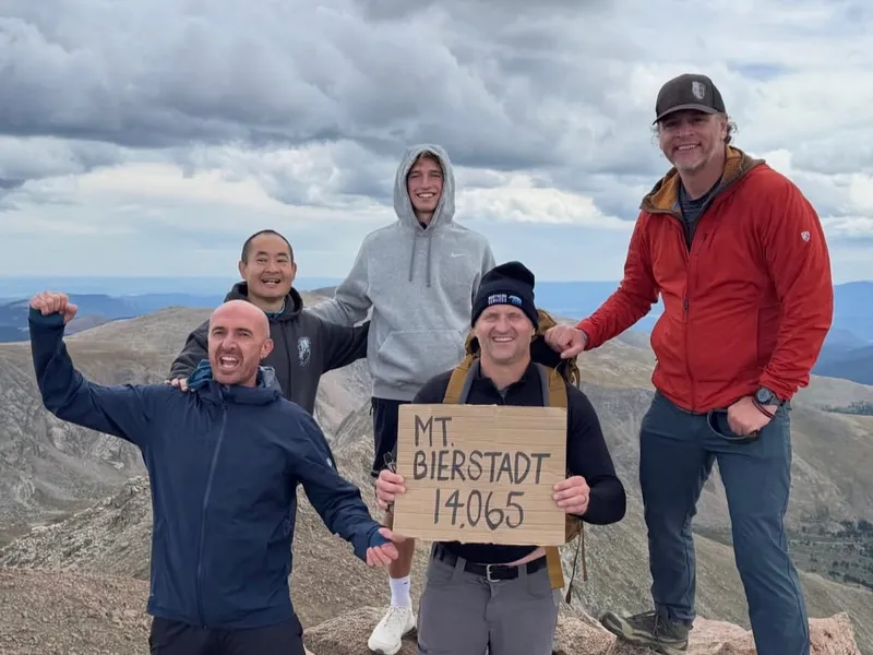 Five hikers on the summit of Mt Bierstadt holding a cardboard sign reading Mt. Bierstadt 14,065