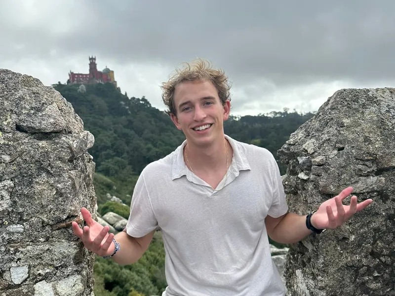Roanin smiling with arms out between old stone battlements, a red and yellow palace on the hill behind