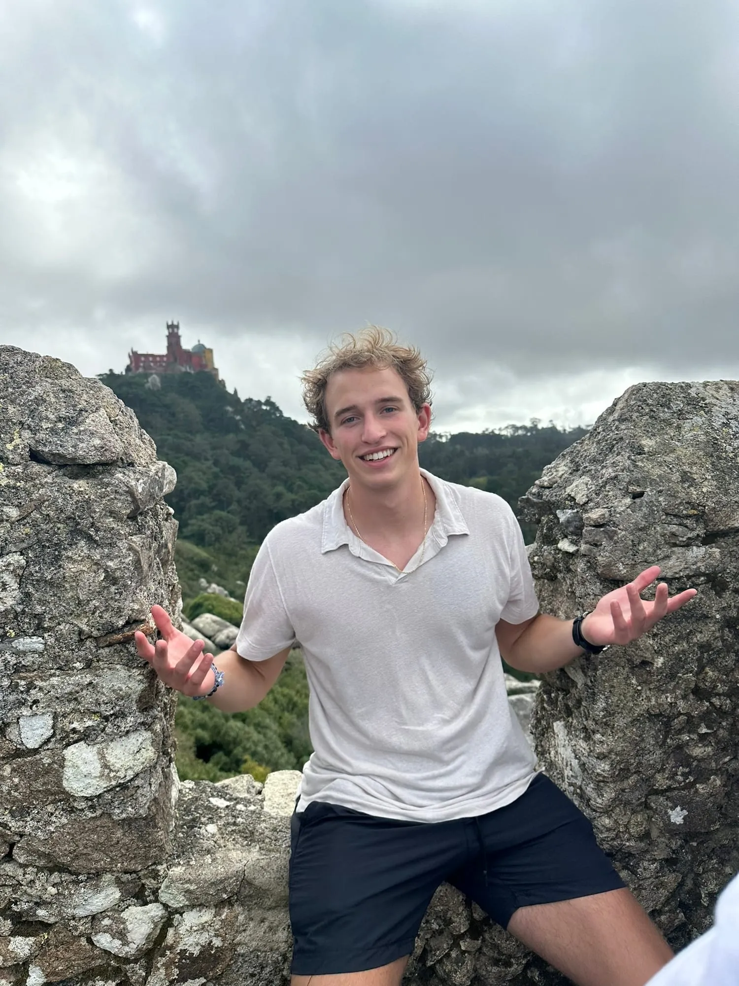 Roanin smiling with arms out between old stone battlements, a red and yellow palace on the hill behind